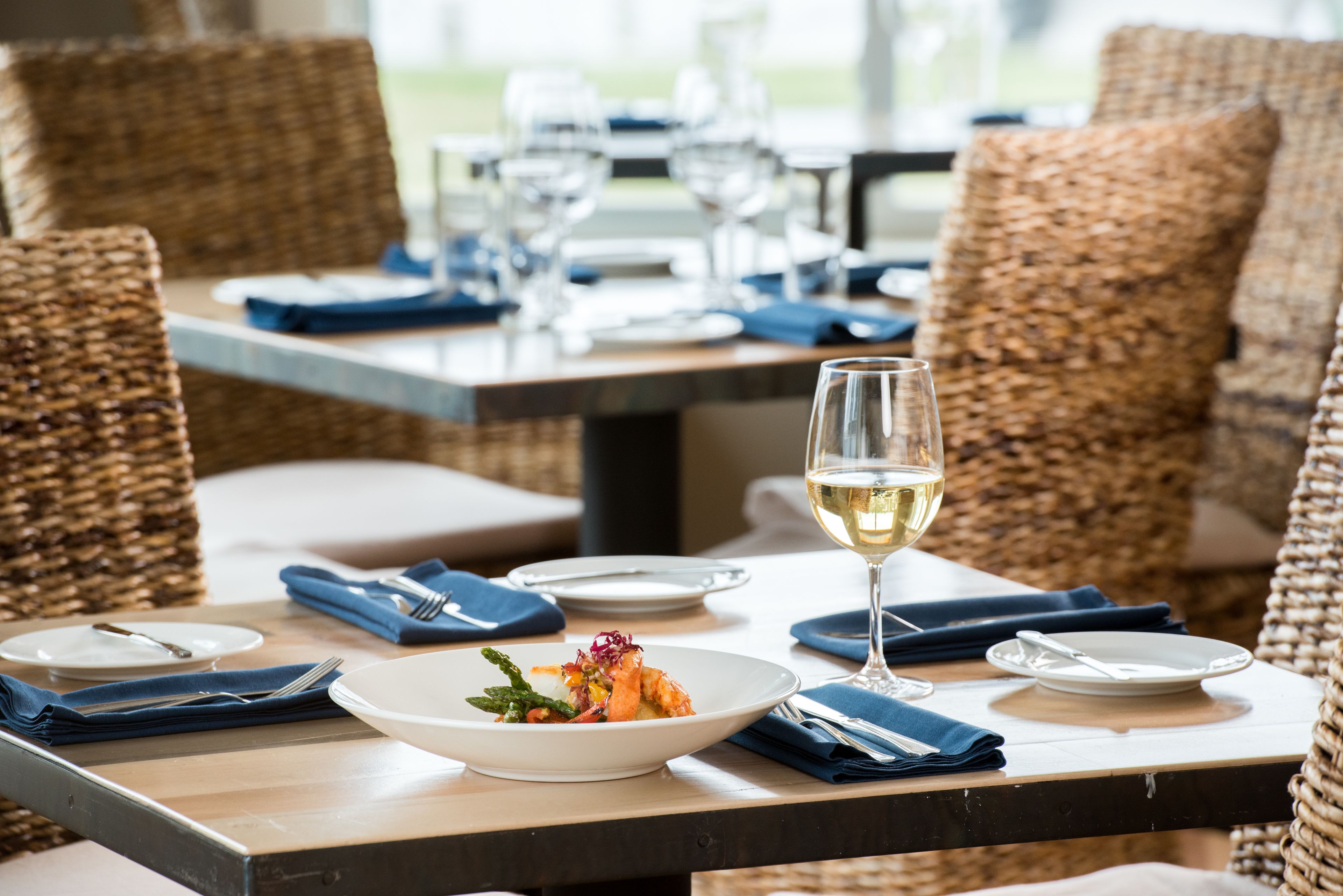 A dining table set with plates, cutlery, and a glass of white wine, surrounded by wicker chairs.