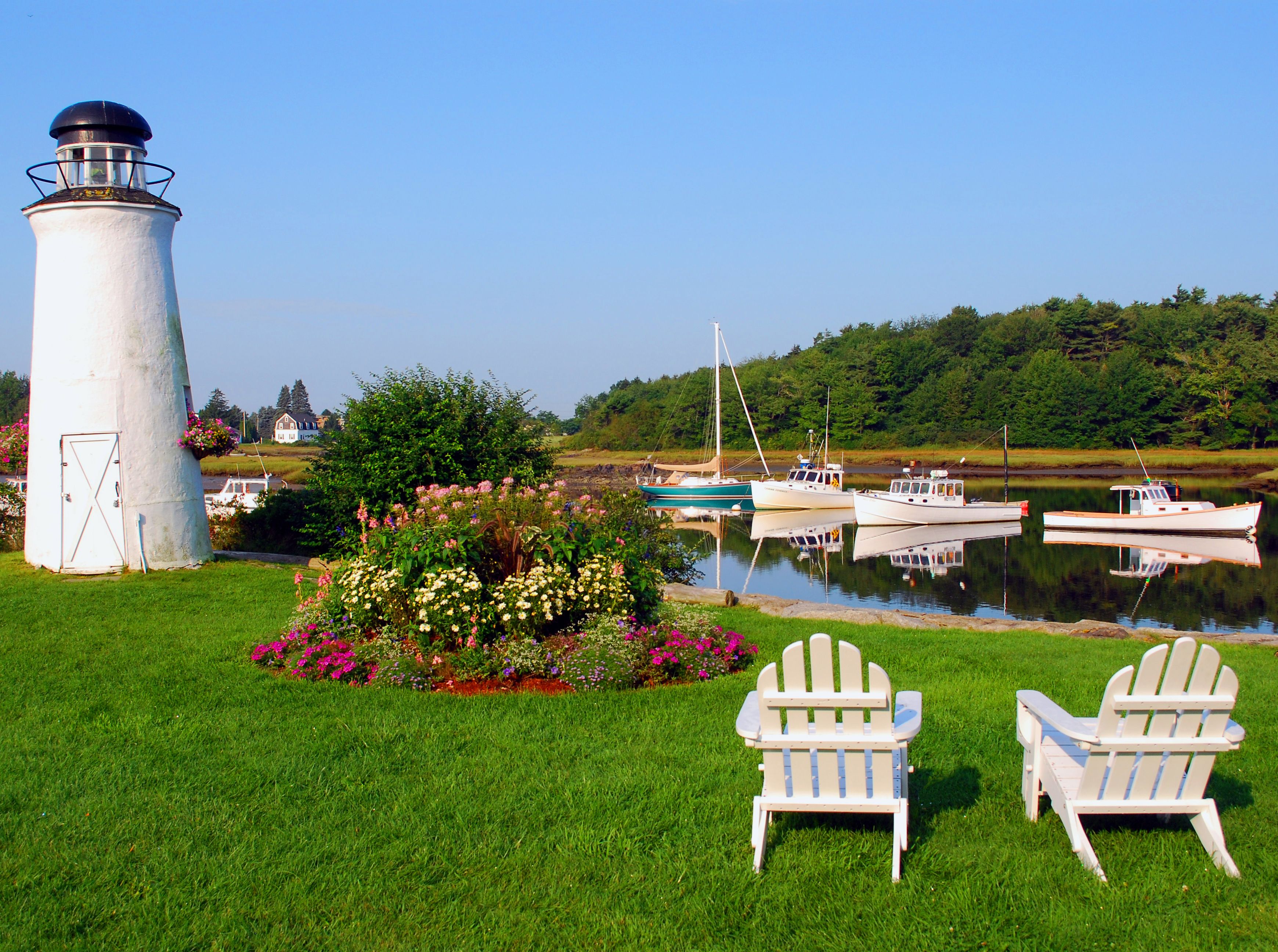 A lighthouse, several boats, and two Adirondack chairs in a grassy area with a lake and trees in the background.