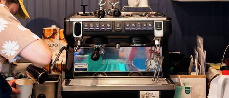 A coffee shop counter with a coffee machine, a menu board, and a shelf with various items.