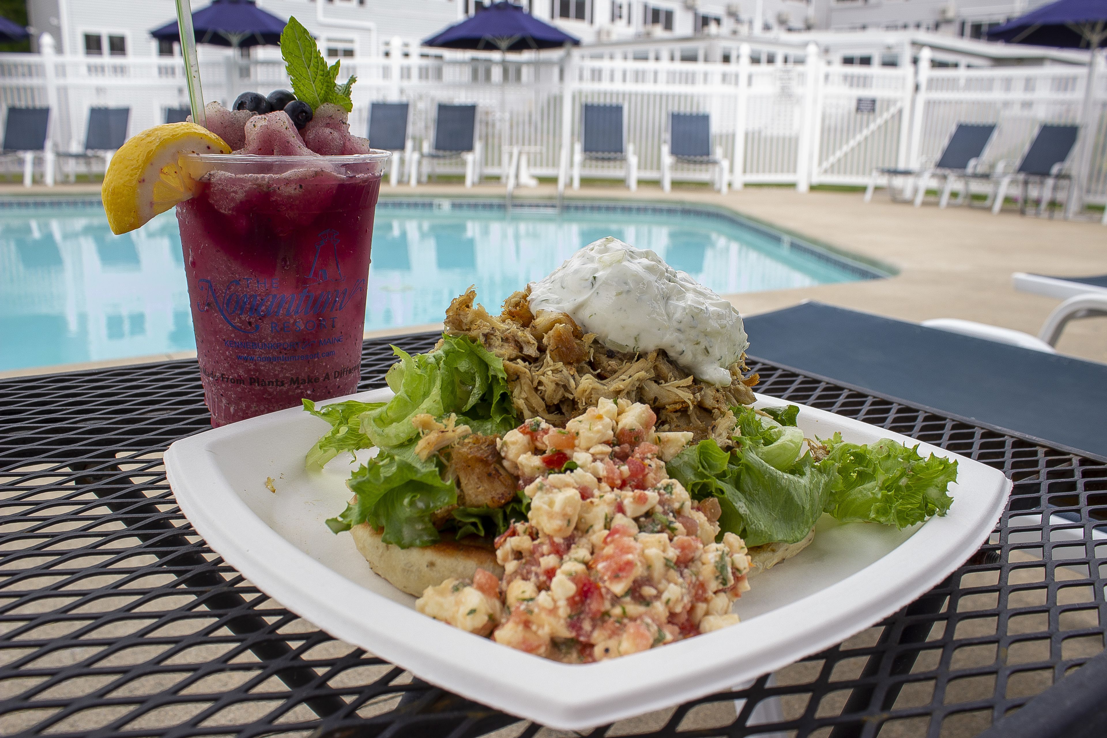 A meal with a salad, chicken, and a drink with a lemon garnish, set on a table by a pool with lounge chairs and umbrellas in the background.