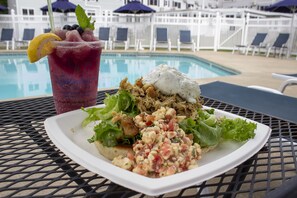 A meal with a salad, chicken, and a drink with a lemon garnish, set on a table by a pool with lounge chairs and umbrellas in the background.