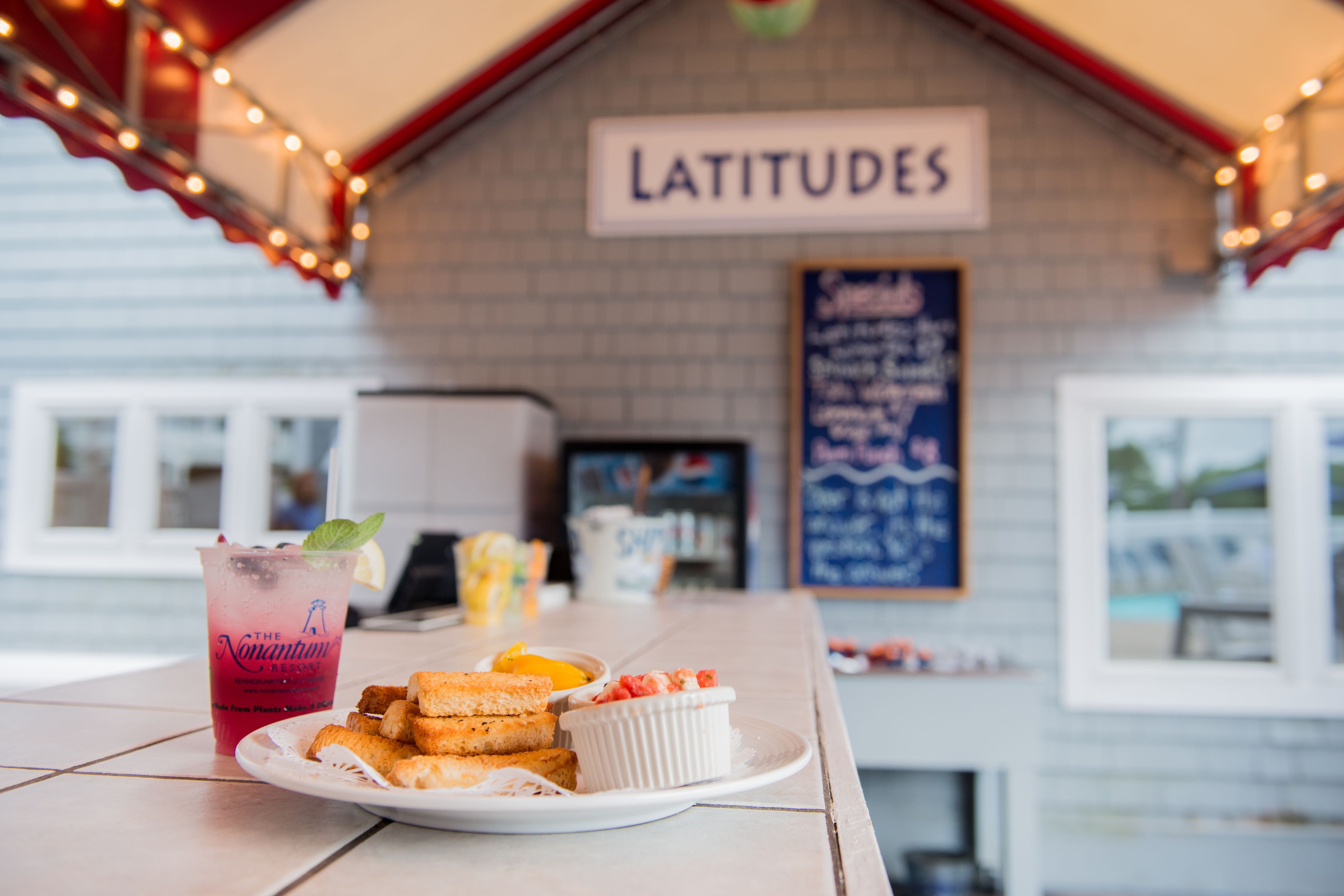 A table with a drink, bread, and a small dish, under a sign reading "LATITUDES".