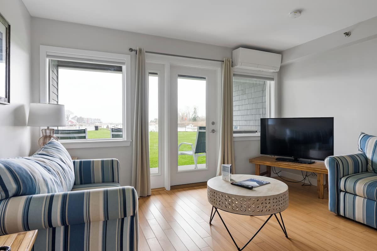 Ett modernt vardagsrum med en soffa, ett soffbord, en tv och stora fönster med gardiner.