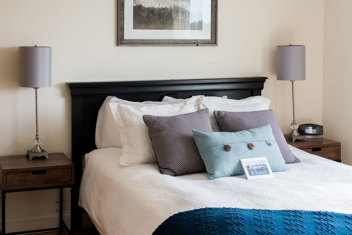 Un lit bien fait, recouvert d’une couvre-lit blanche et bleue, avec deux coussins à motifs et un tableau encadré au-dessus.
