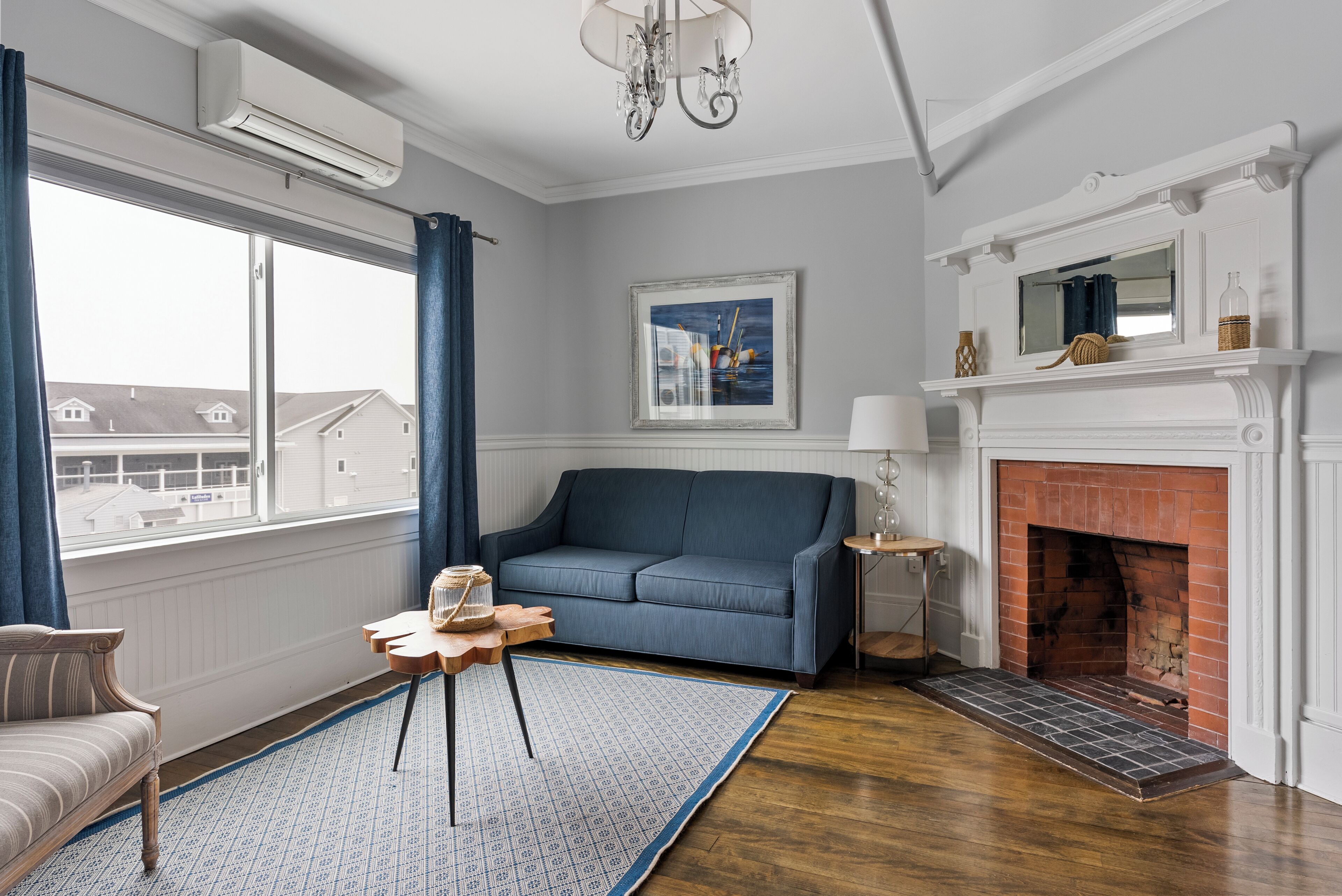 A living room with a blue sofa, a small wooden table, a fireplace, and a view of houses through a large window.