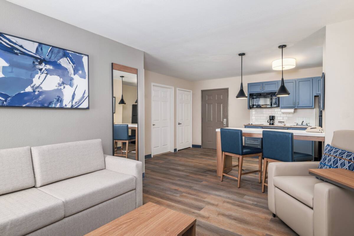 A modern living room with a sofa, a coffee table, and a dining area with chairs and a kitchen in the background.