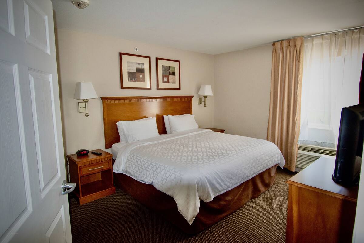 A hotel room with a large bed, two bedside tables, a TV, and a window with curtains.