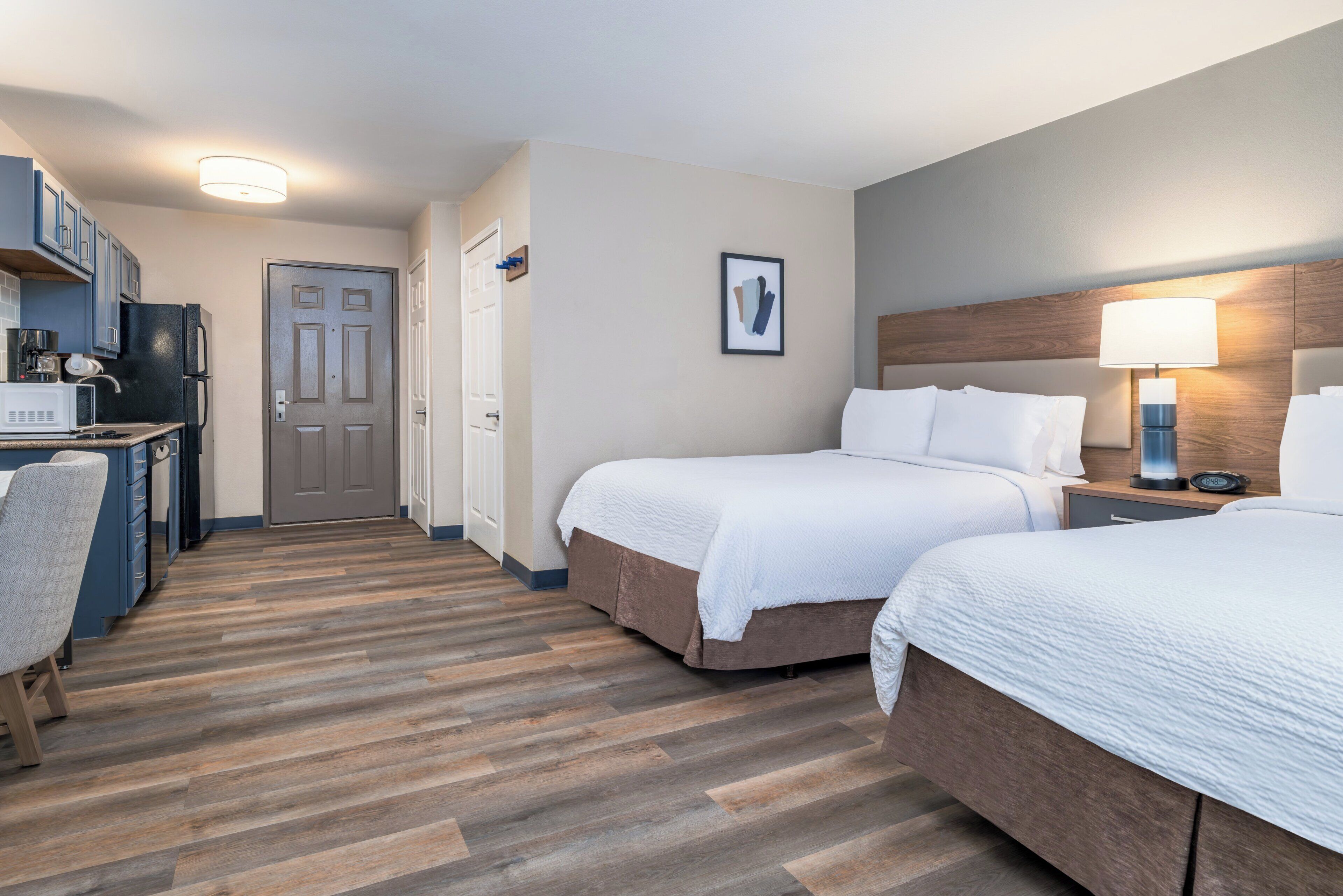 Een hotelkamer met twee bedden, een kitchenette, een deur en een lijst met een afbeelding aan de muur.