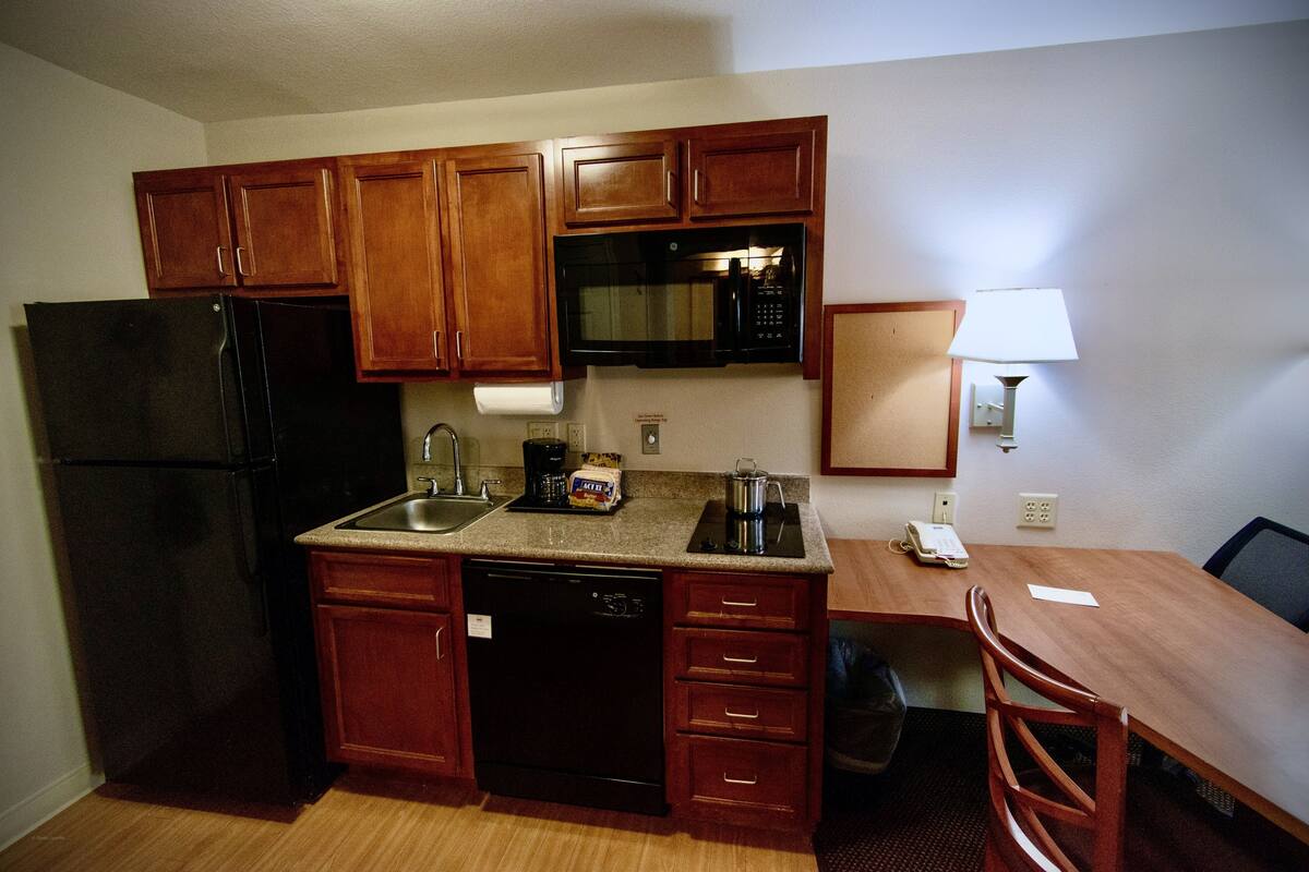 A compact kitchen with wooden cabinets, a black refrigerator, a microwave, a coffee maker, and a small table with a chair.