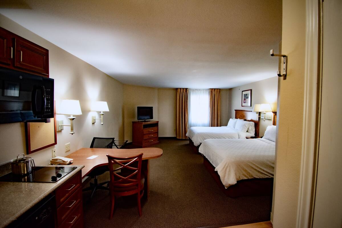 A hotel room with a kitchenette, a bed, a desk with a chair, and two bedside tables with lamps.