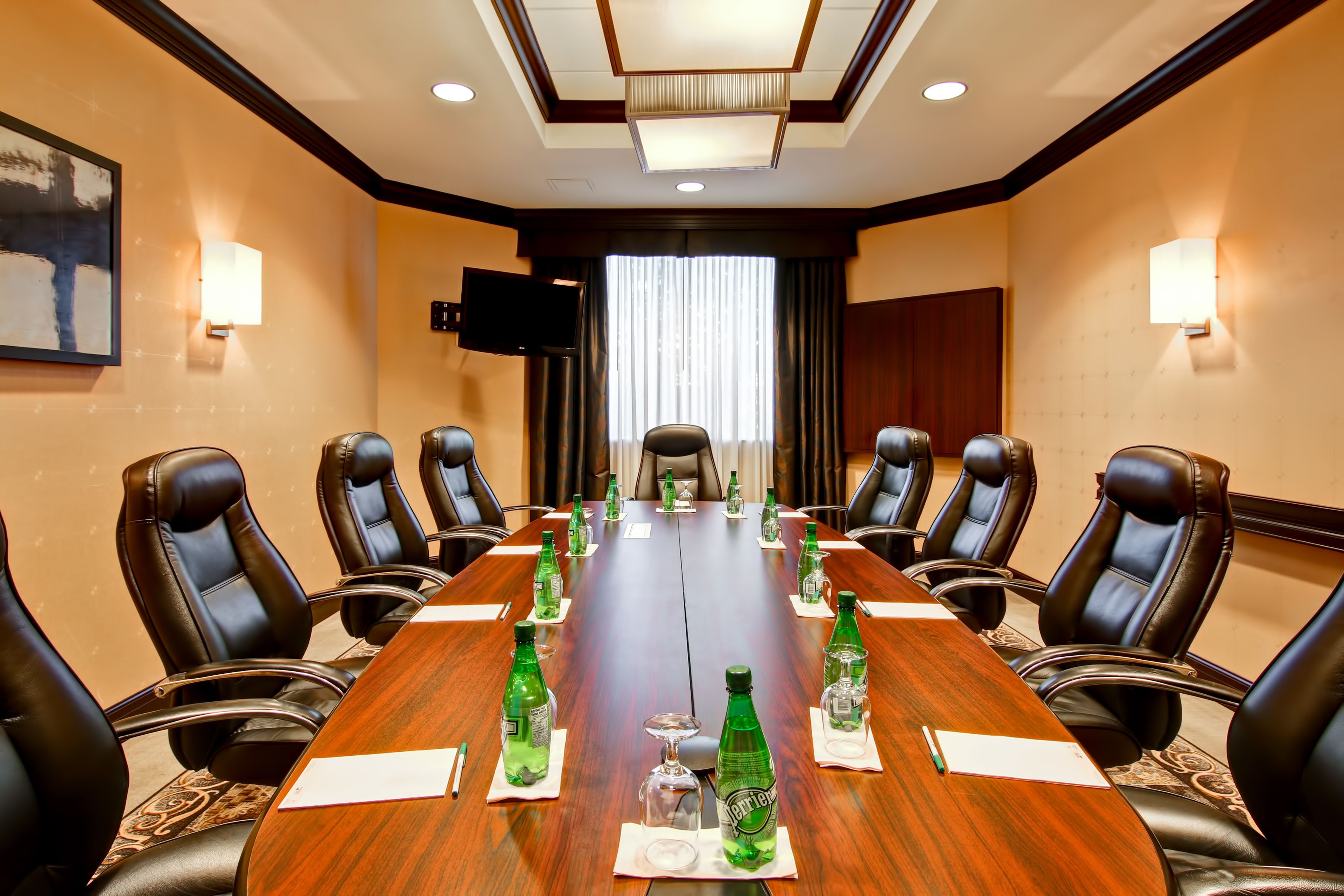 A conference room with a long wooden table, black leather chairs, and a television on the wall.