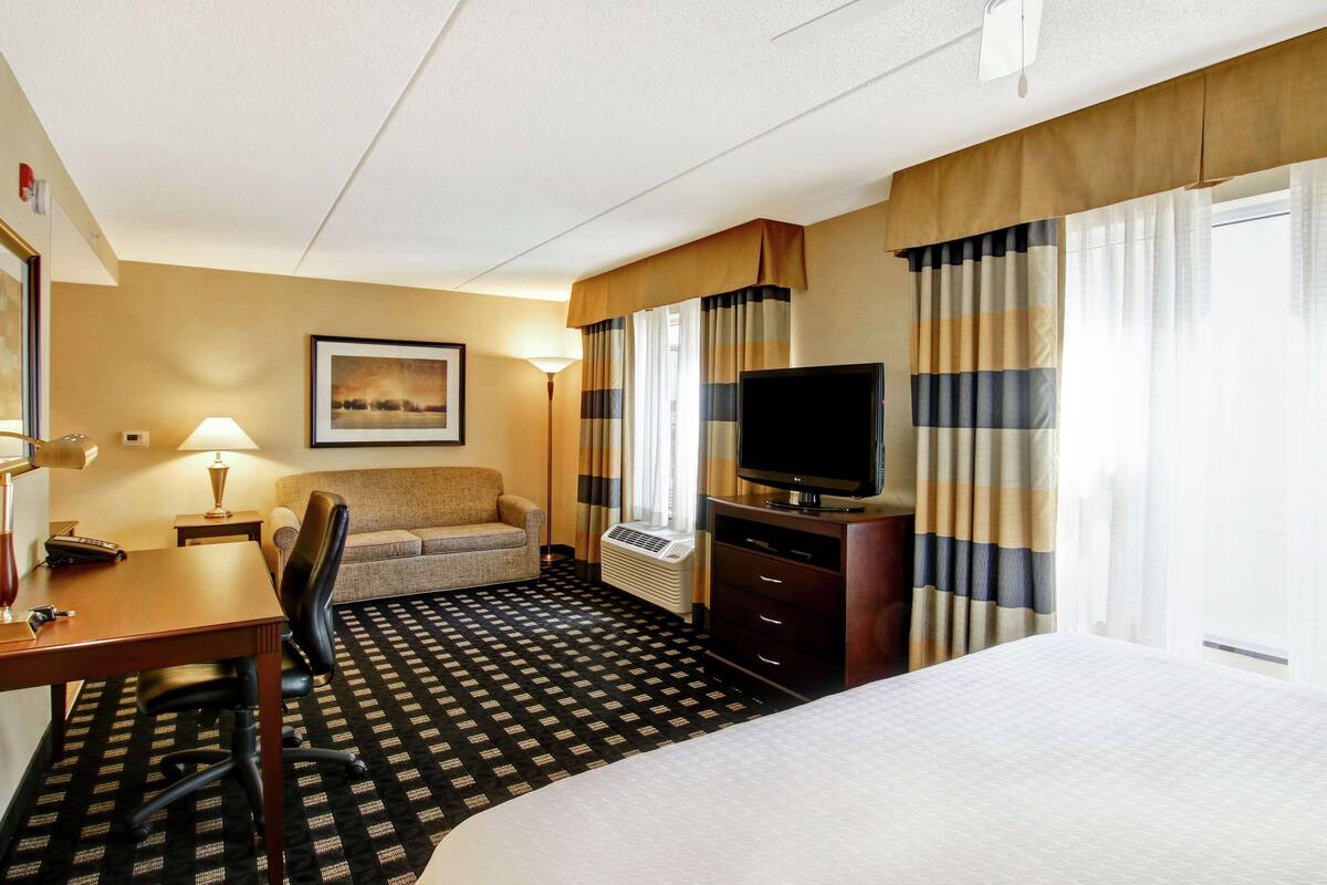 A hotel room with a bed, a desk with a chair, a television, a lamp, and a framed picture on the wall.