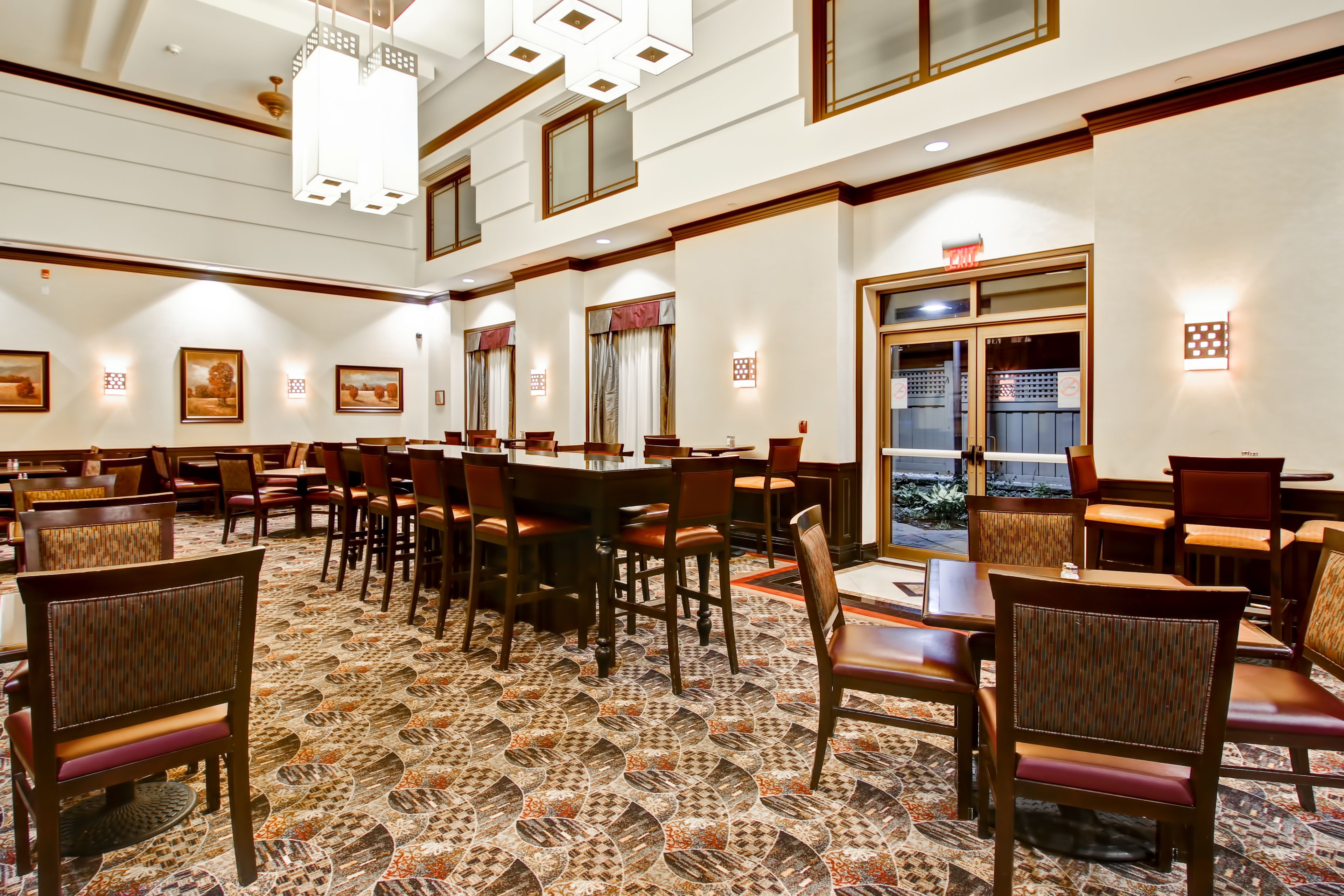 A spacious hotel lobby with multiple dining tables and chairs, a patterned carpet, and framed artwork on the walls.