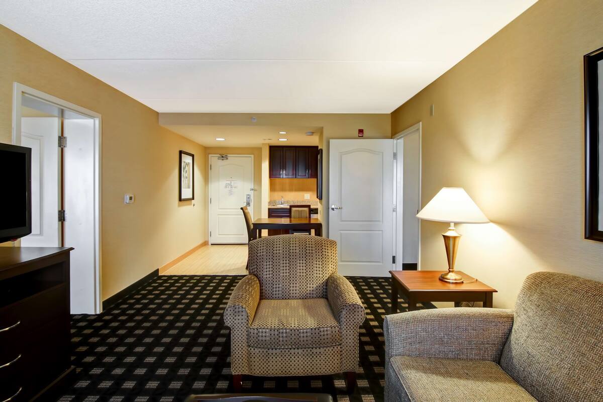 A hotel room with a patterned carpet, a sofa, a chair, a TV, a lamp, and a dining area with a table and chairs.