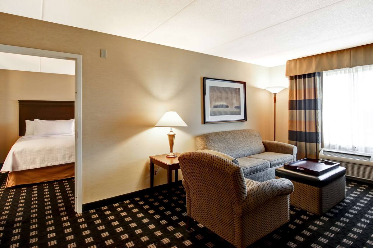 A hotel room with a bed, a sofa, a chair, a lamp, a tray table, and a window with curtains.