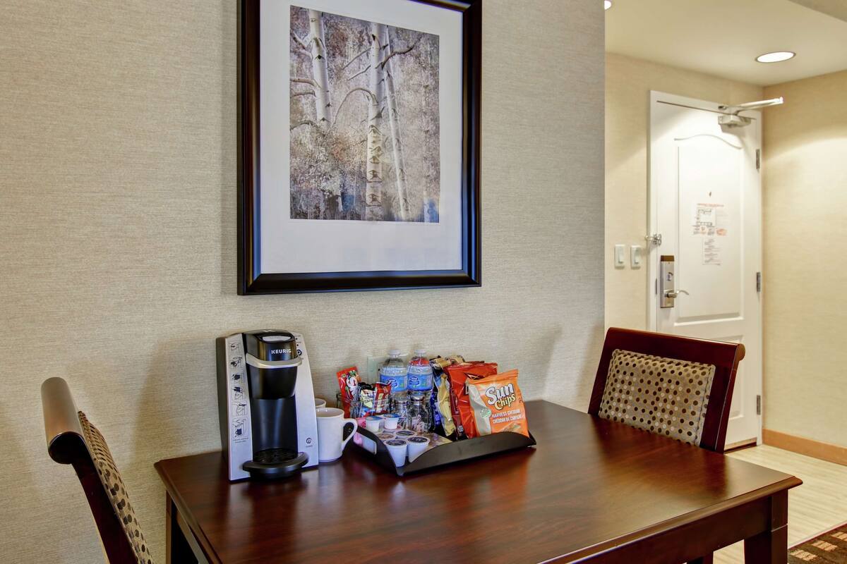 A hotel room with a wooden table, a framed picture on the wall, a coffee maker, and a tray with snacks and bottled water.