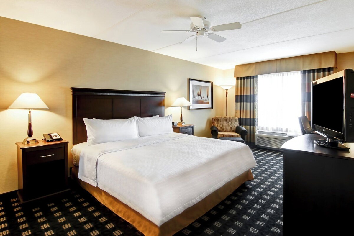 A hotel room with a bed, two bedside tables with lamps, a chair, a desk with a TV, and a ceiling fan.