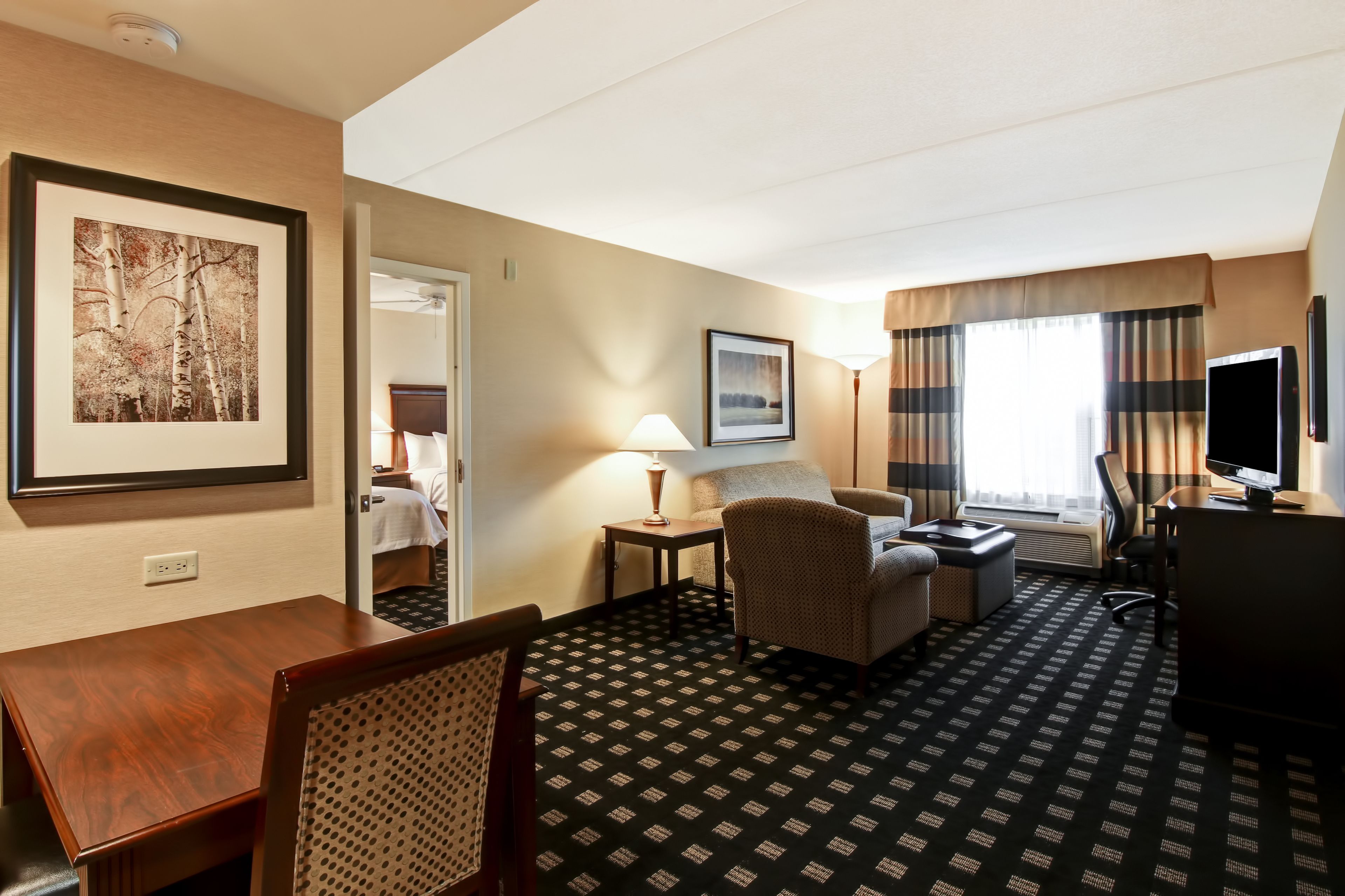A hotel room with a wooden desk, a chair, a sofa, a TV, and a bed in the background.