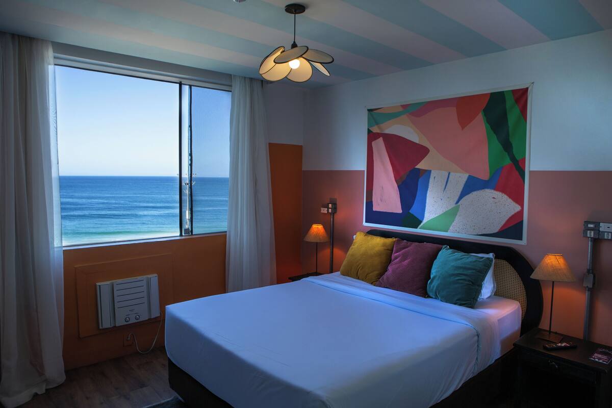 Habitación de hotel con una cama grande, vista al océano, un cuadro abstracto colorido en la pared y dos lámparas de noche.