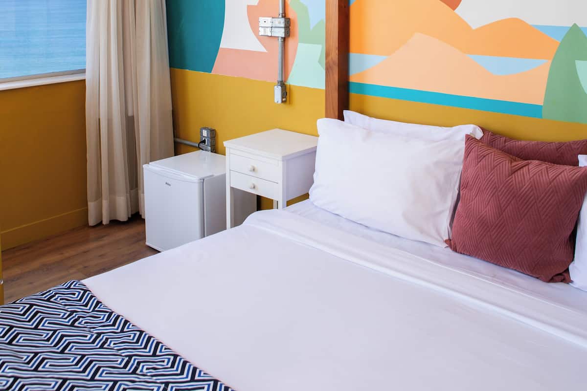 Habitación de hotel con cama, mesitas de noche, una lámpara colgante de bambú, una pared con un mural colorido y una ventana con vistas al mar.