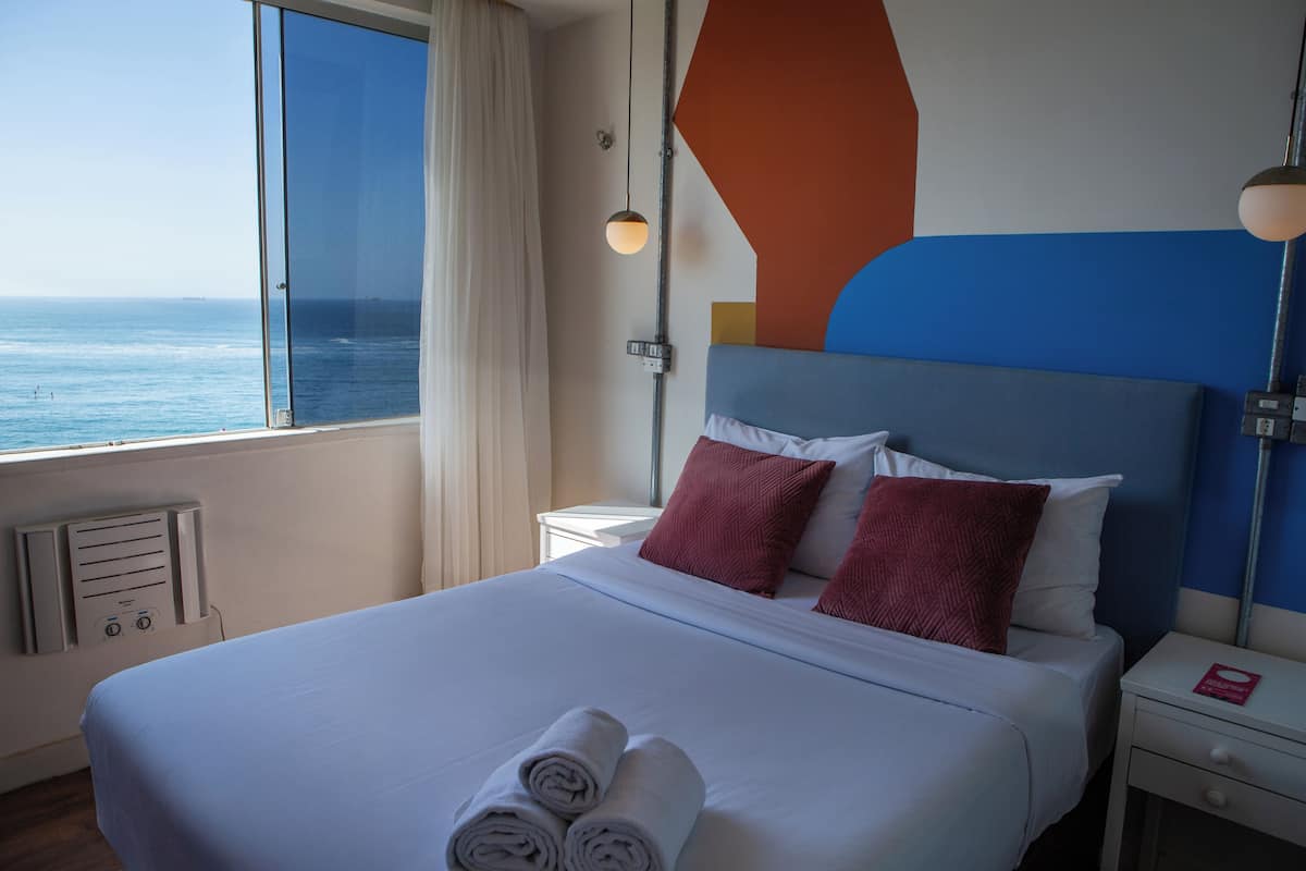 Habitación de hotel con una cama grande, una mesita de noche, una lámpara y vista al océano.