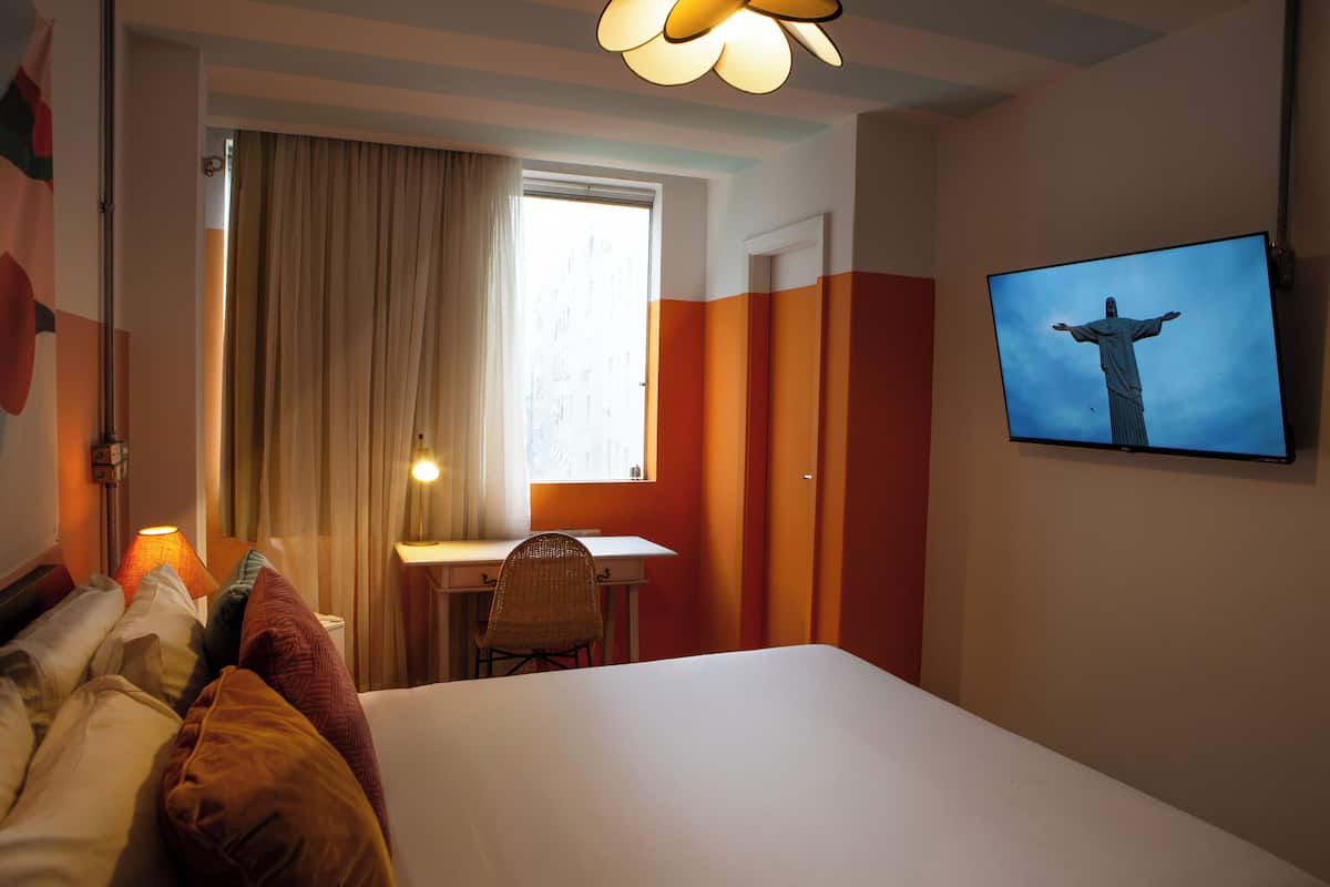 Habitación de hotel con cama, escritorio, silla, televisor mostrando la estatua del Cristo Redentor y ventana con cortinas.