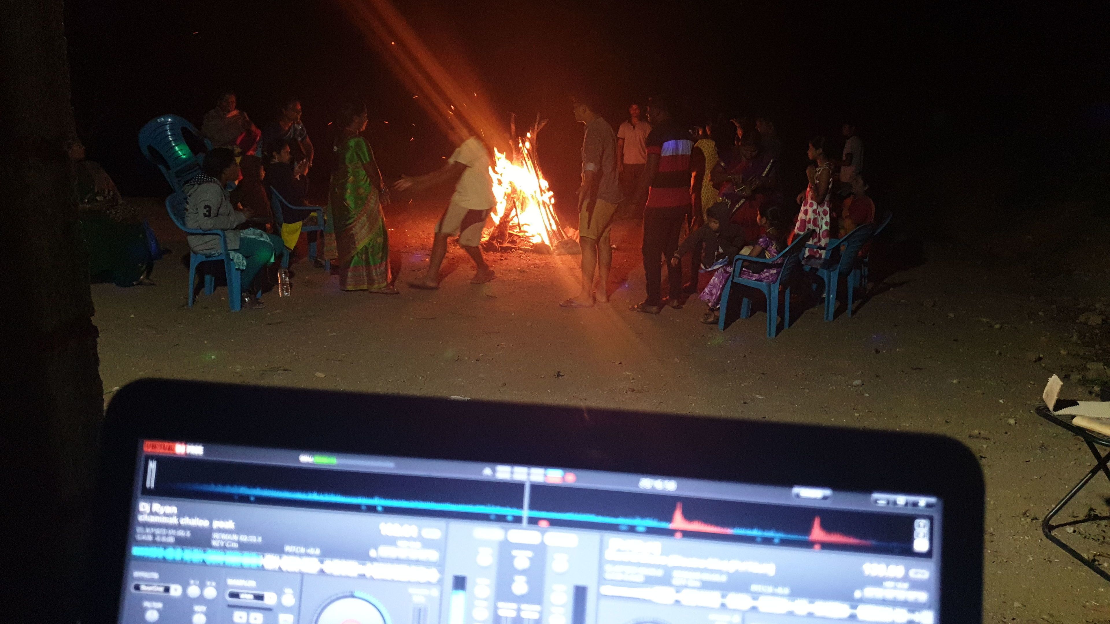 A group of people gathered around a campfire at night, with a laptop displaying DJ software in the foreground.