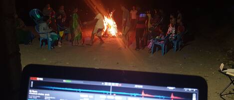 A group of people gathered around a campfire at night, with a laptop displaying DJ software in the foreground.