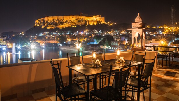 A terrace with tables and chairs overlooking a cityscape with a prominent hilltop structure. - Jee Ri Haveli (Jodhpur)