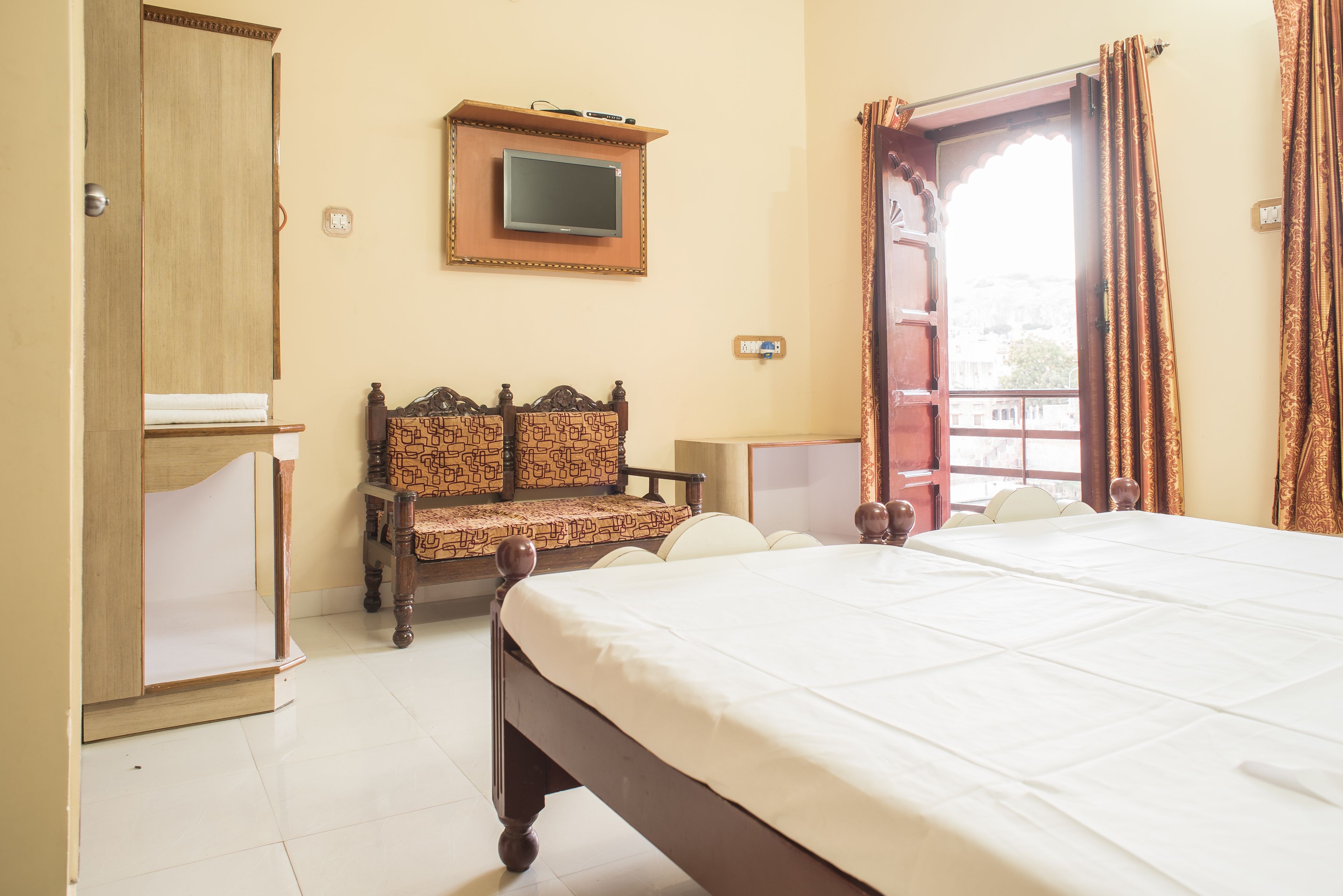 A hotel room with a bed, a wooden wardrobe, a television mounted on the wall, and a window with curtains.