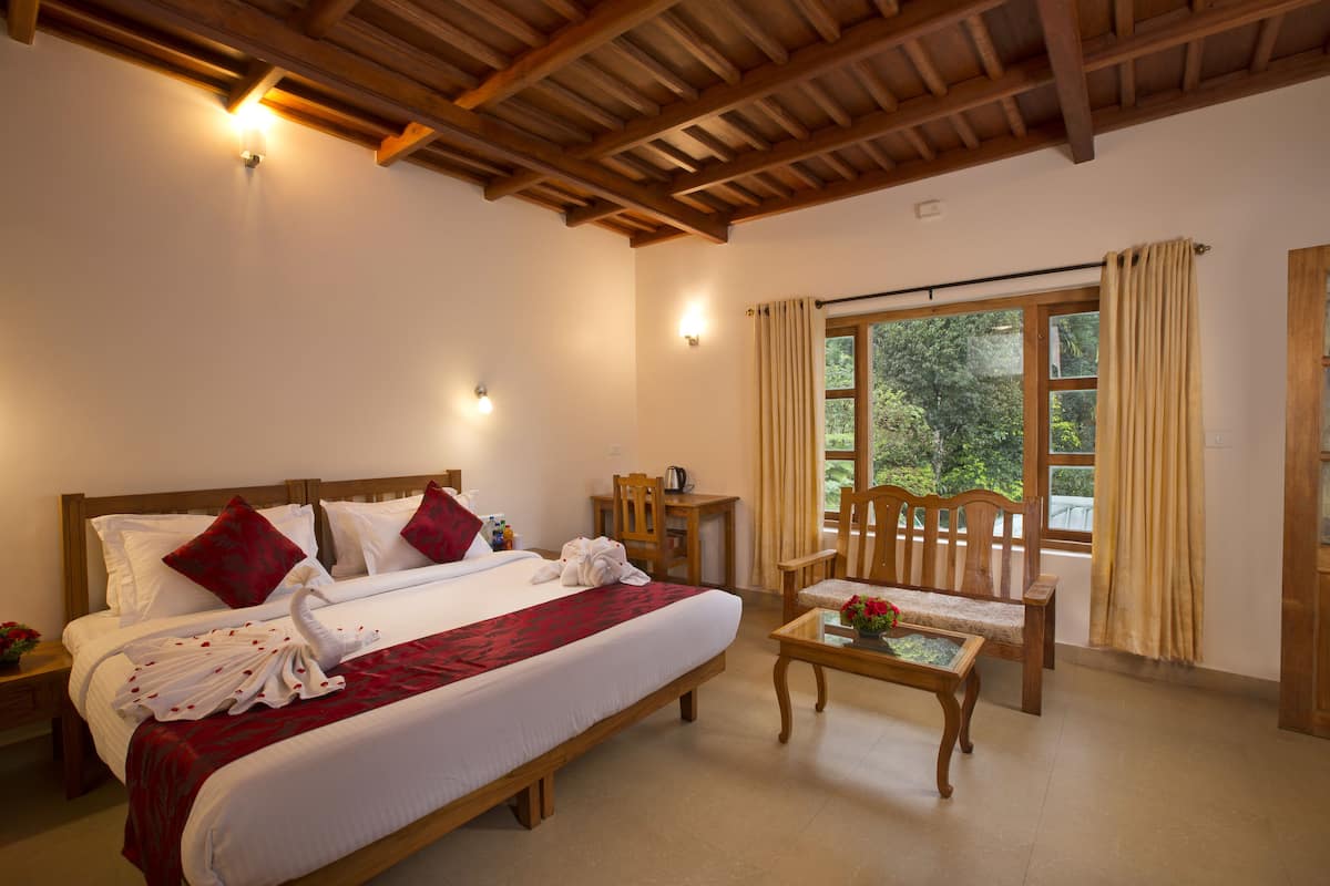 A hotel room with a large bed, wooden headboard, two bedside tables, a wooden chair, a small table with a vase of flowers, and a window with curtains.