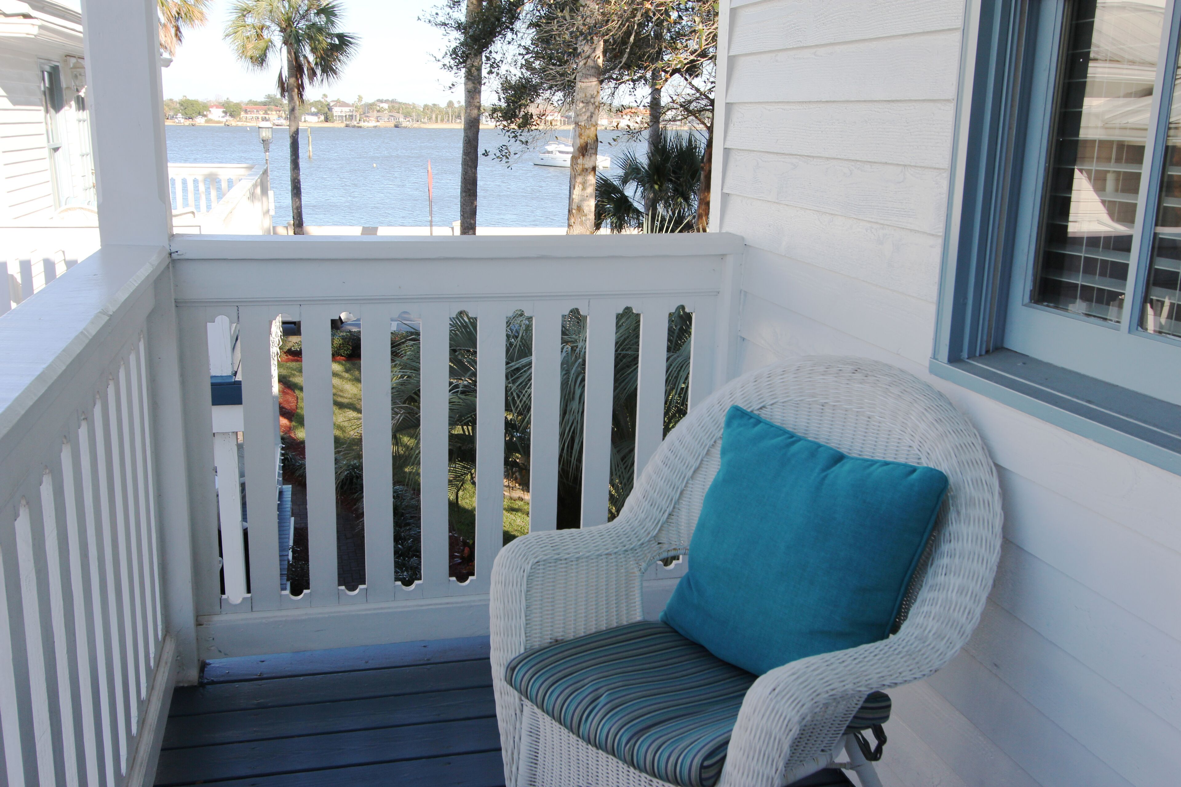A white wicker chair with a blue cushion on a porch with a white railing, overlooking a body of water and trees.