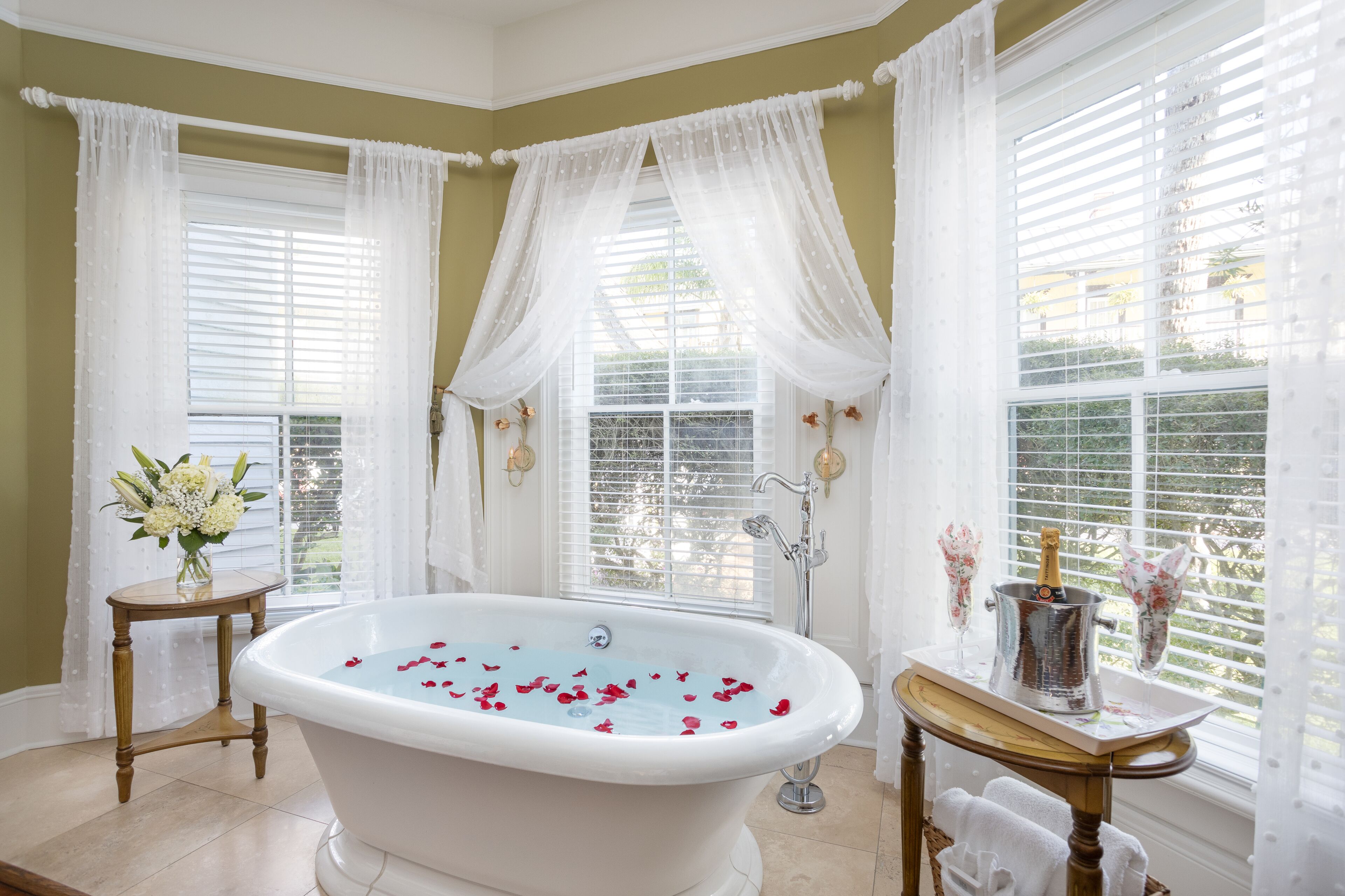 A bathroom with a freestanding bathtub, white curtains, and a vase of flowers on a side table.