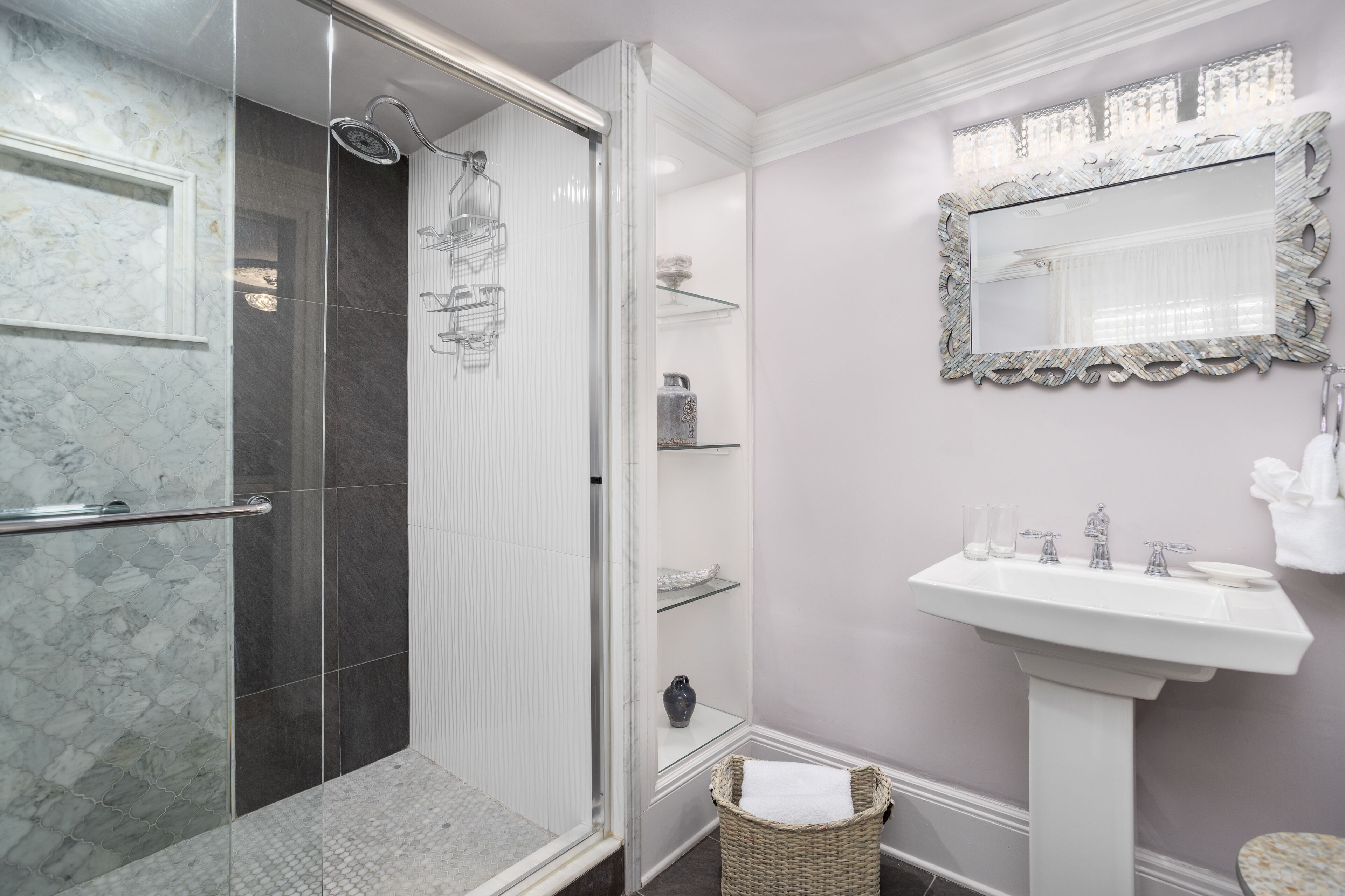 A bathroom with a glass-enclosed shower, a white pedestal sink, and a mirror with a decorative frame.