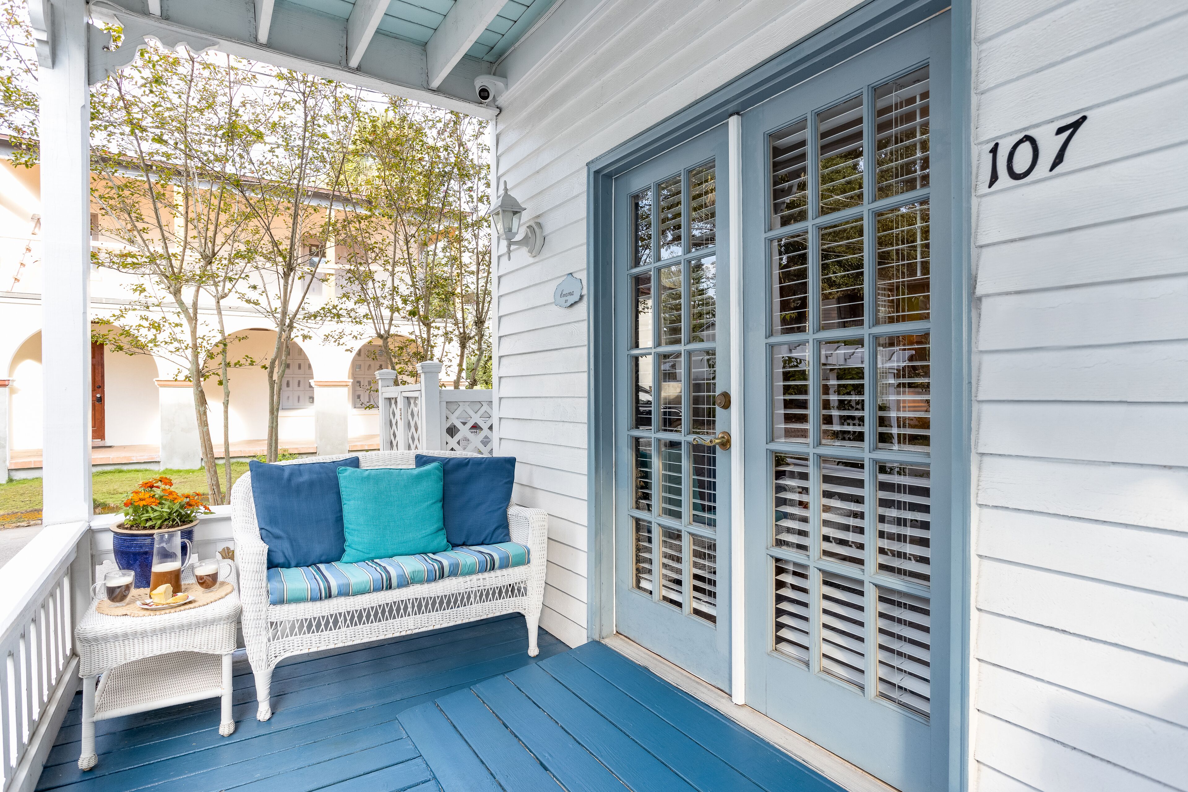 A porch with a white wicker sofa, a small table with drinks, and a blue door with the number 107.