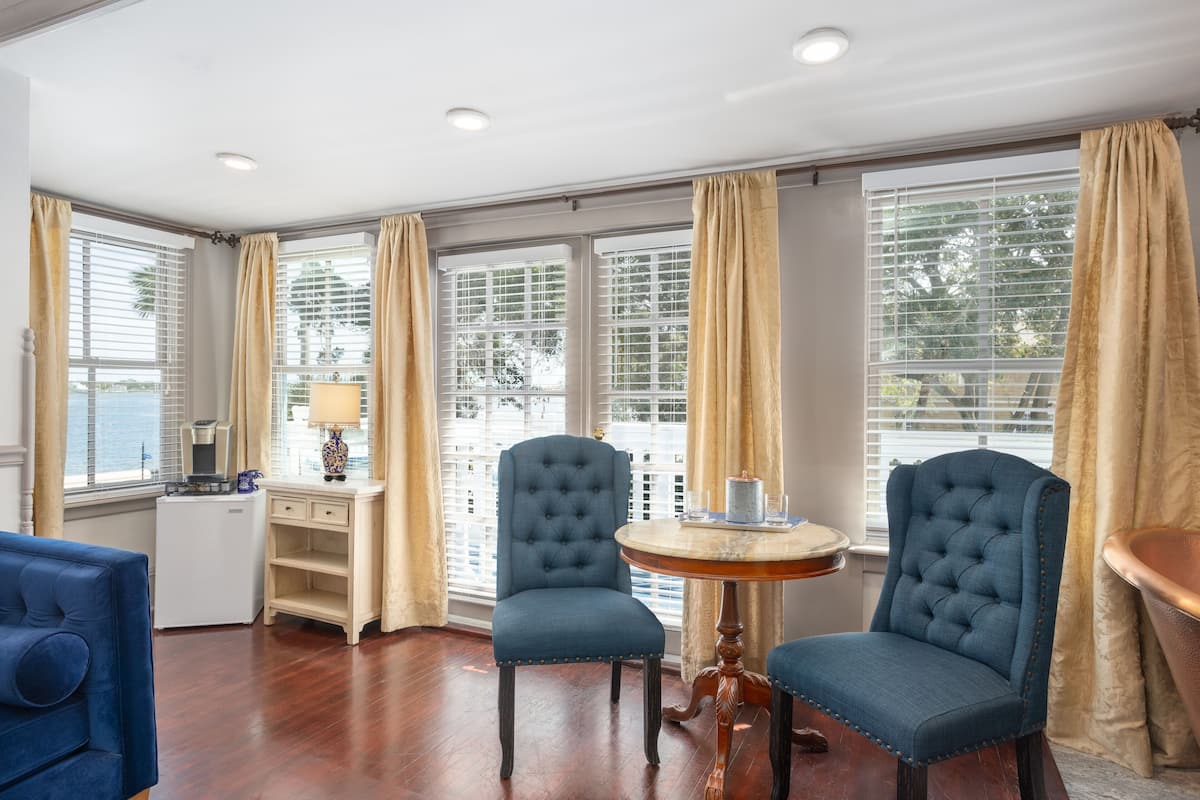 Una habitación con dos sillones tapizados en azul, una mesita redonda, un escritorio blanco con lámpara y vistas amplias a través de ventanas con persianas.