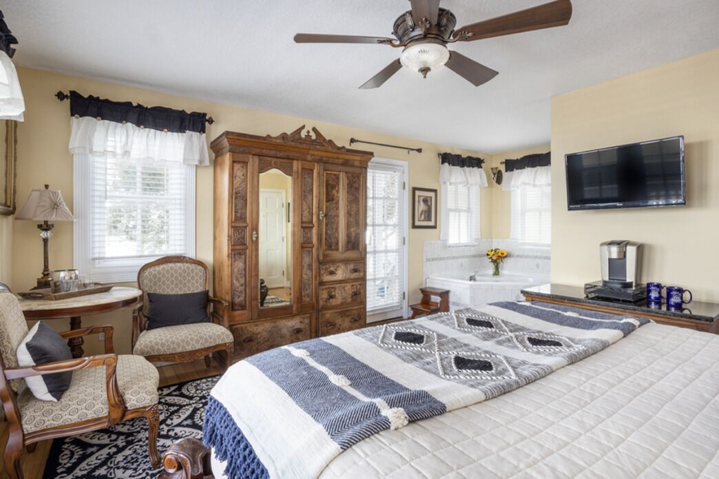 A bedroom with a bed, a wooden wardrobe, a desk with a lamp, a television, and a ceiling fan.