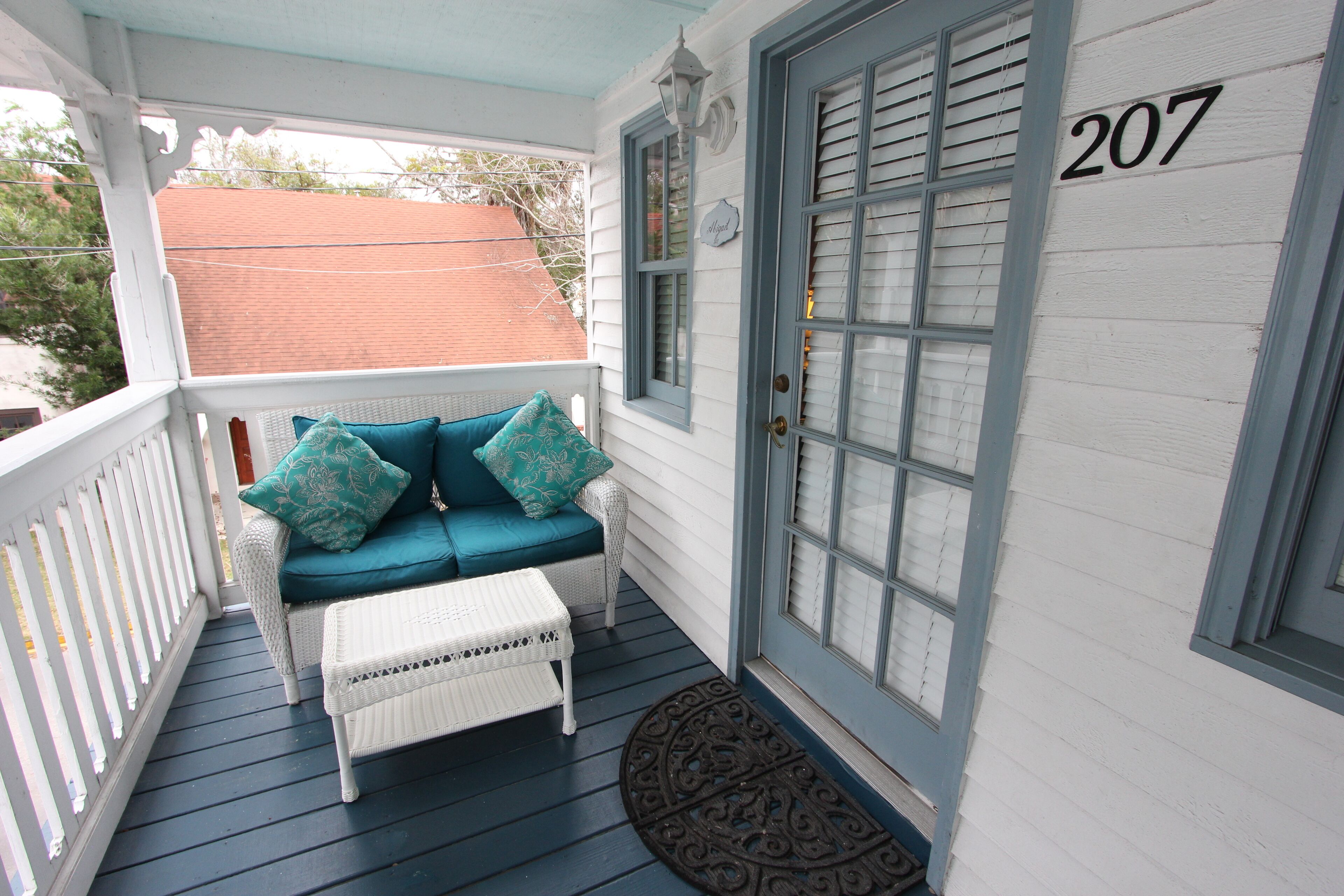 A porch with a white wicker sofa and teal cushions, a white wicker table, and a door with the number 207.