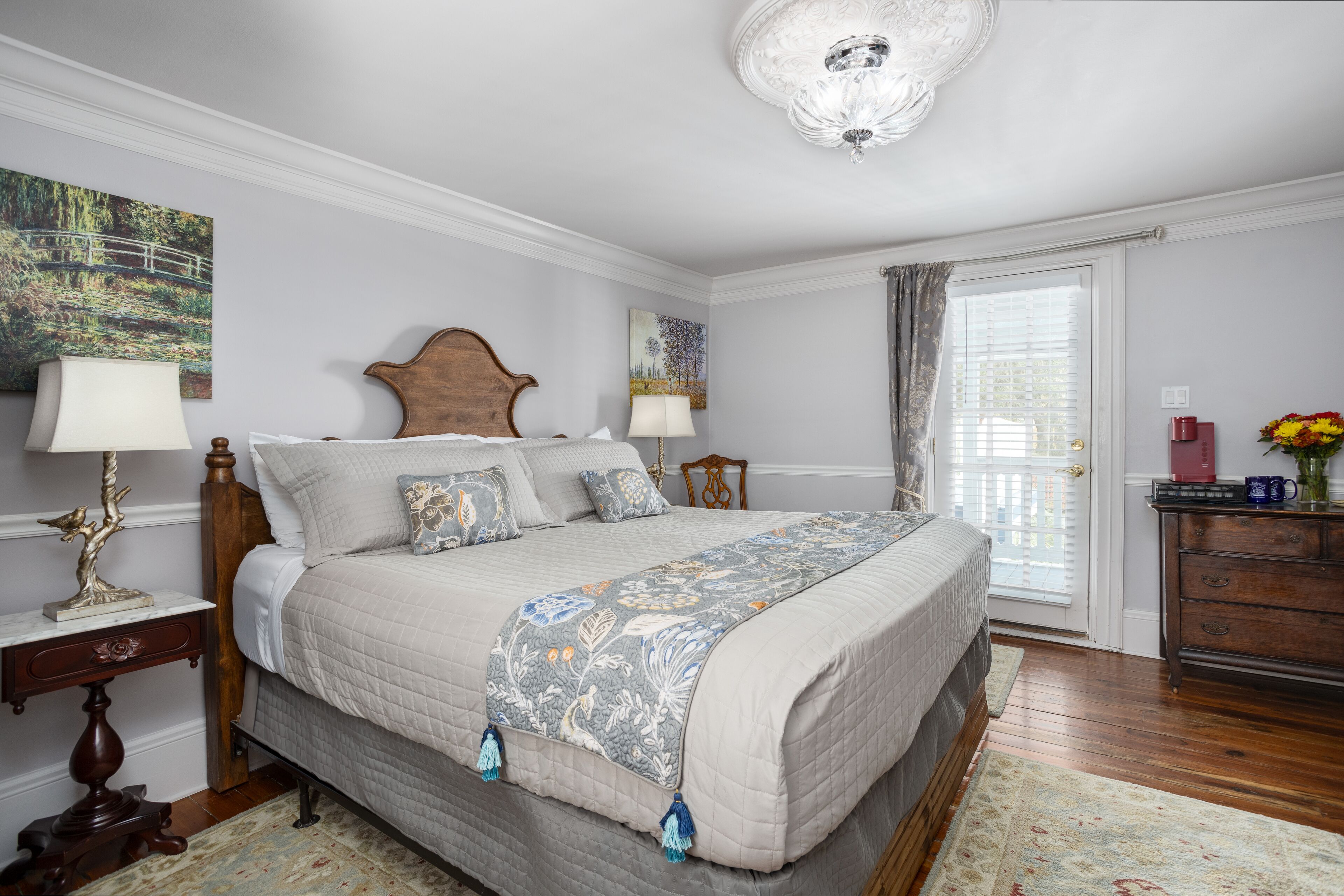 A bedroom with a large bed, two bedside tables with lamps, a wooden headboard, a chandelier, a window with curtains, and a dresser with a vase of flowers.