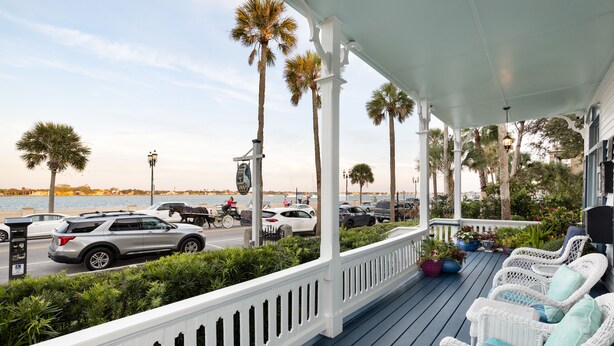 A covered porch with wicker furniture and potted plants, overlooking a street with parked cars and palm trees. - Bayfront Westcott House St Augustine B&B (St. Augustine)