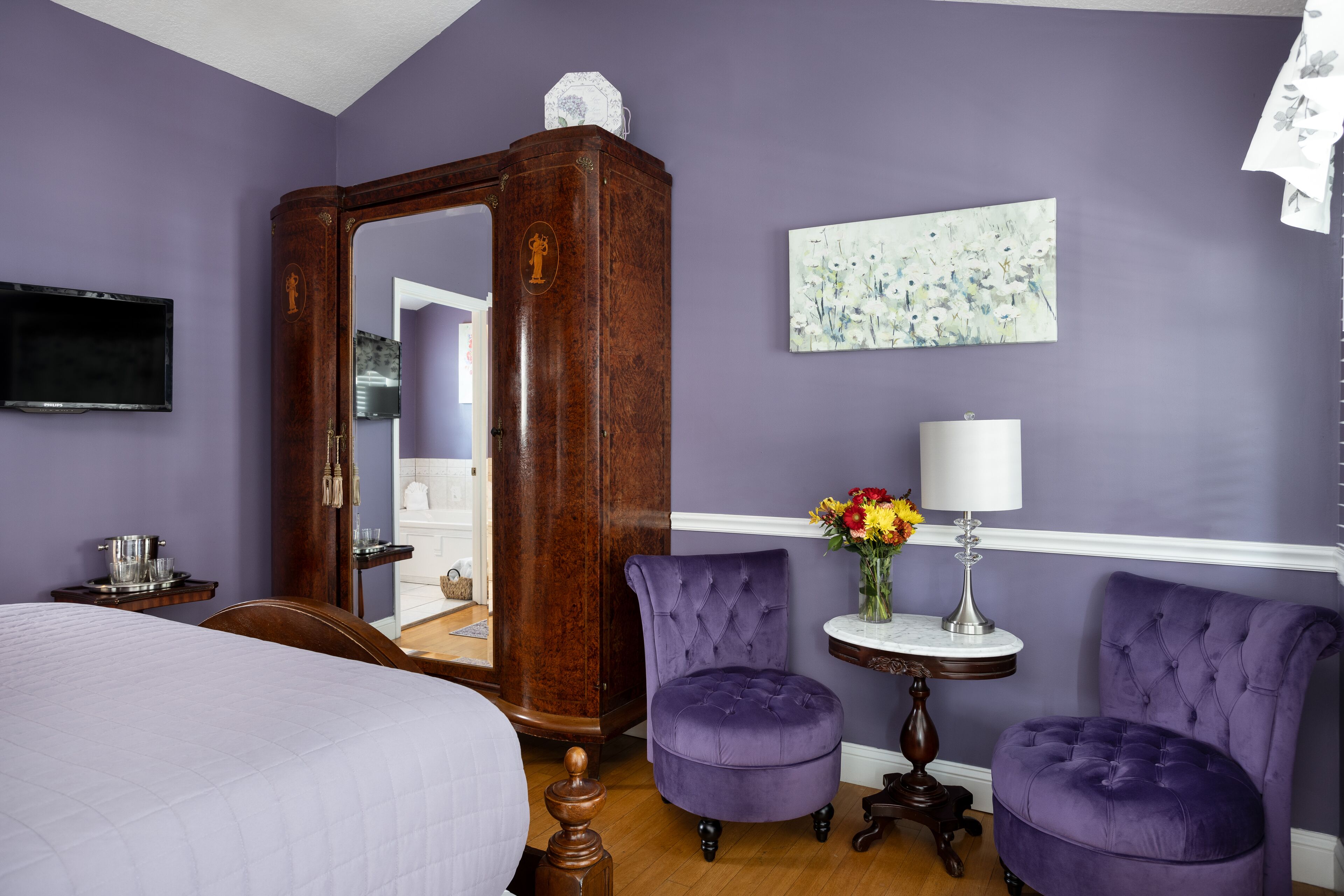 A room with a large mirror, two purple armchairs, a bed, a small table with a lamp, and a painting on the wall.