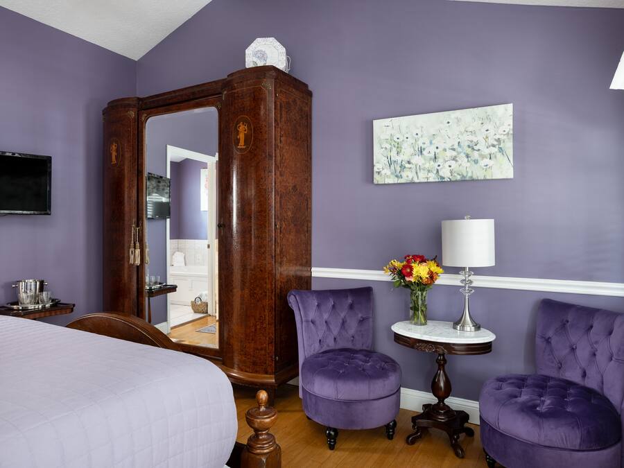 A room with a large mirror, two purple armchairs, a bed, a small table with a lamp, and a painting on the wall.
