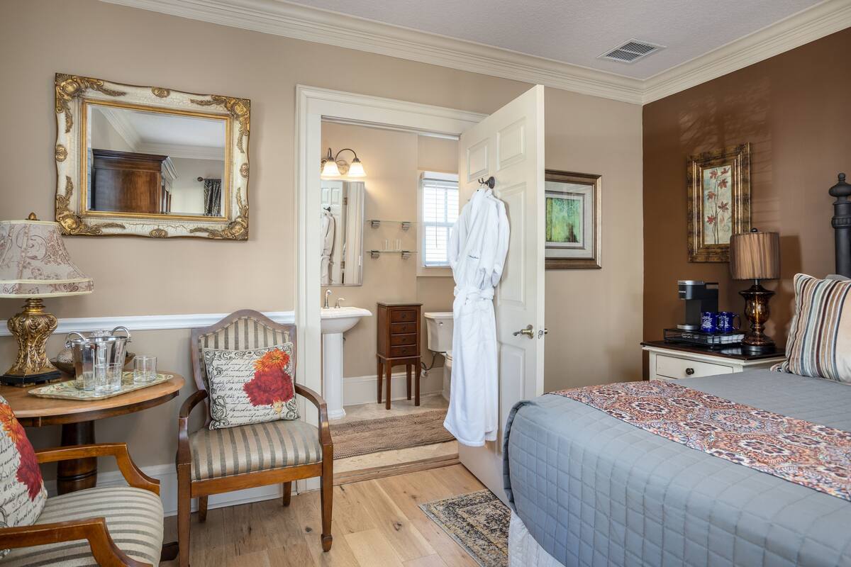 Habitación de hotel con cama, una silla, una mesa con lámpara, un espejo, baño con lavamanos y bata colgada, y mesita de noche con lámpara y cafetera.