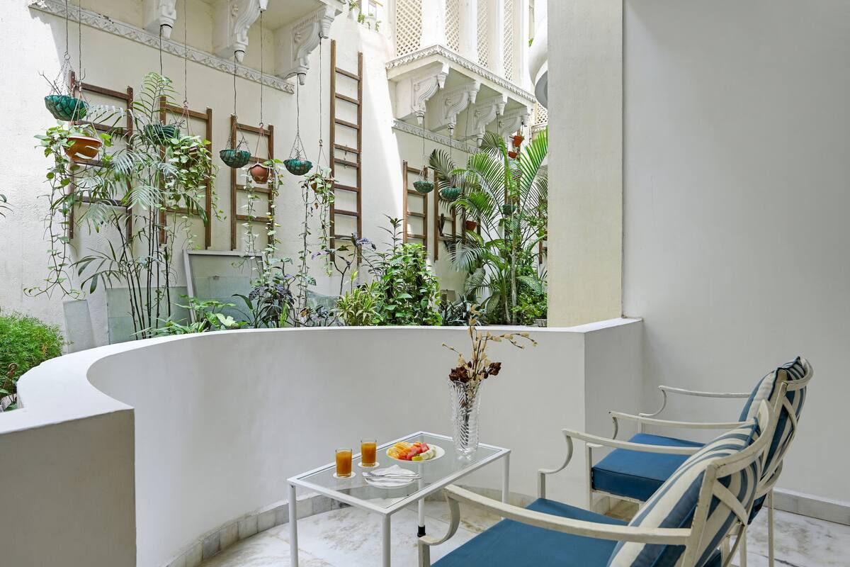 Un balcon avec une table dressée pour le petit-déjeuner, entouré d’une végétation luxuriante et agrémenté de détails architecturaux blancs.