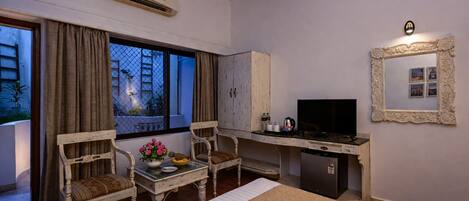 A hotel room with a bed, a desk with a TV, a chair, a small table with flowers, and a window with curtains.