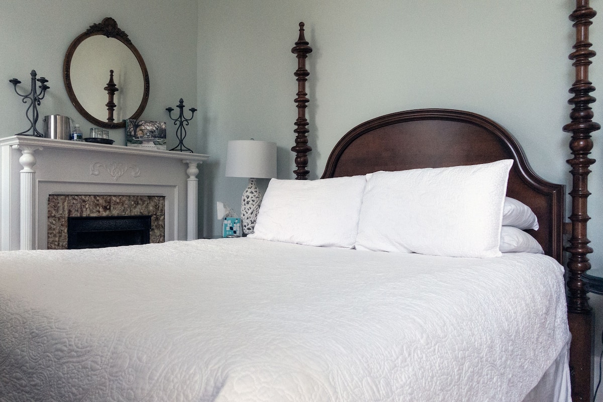 A neatly made bed with white linens, a wooden headboard, and a white wall with a fireplace and a mirror above it.