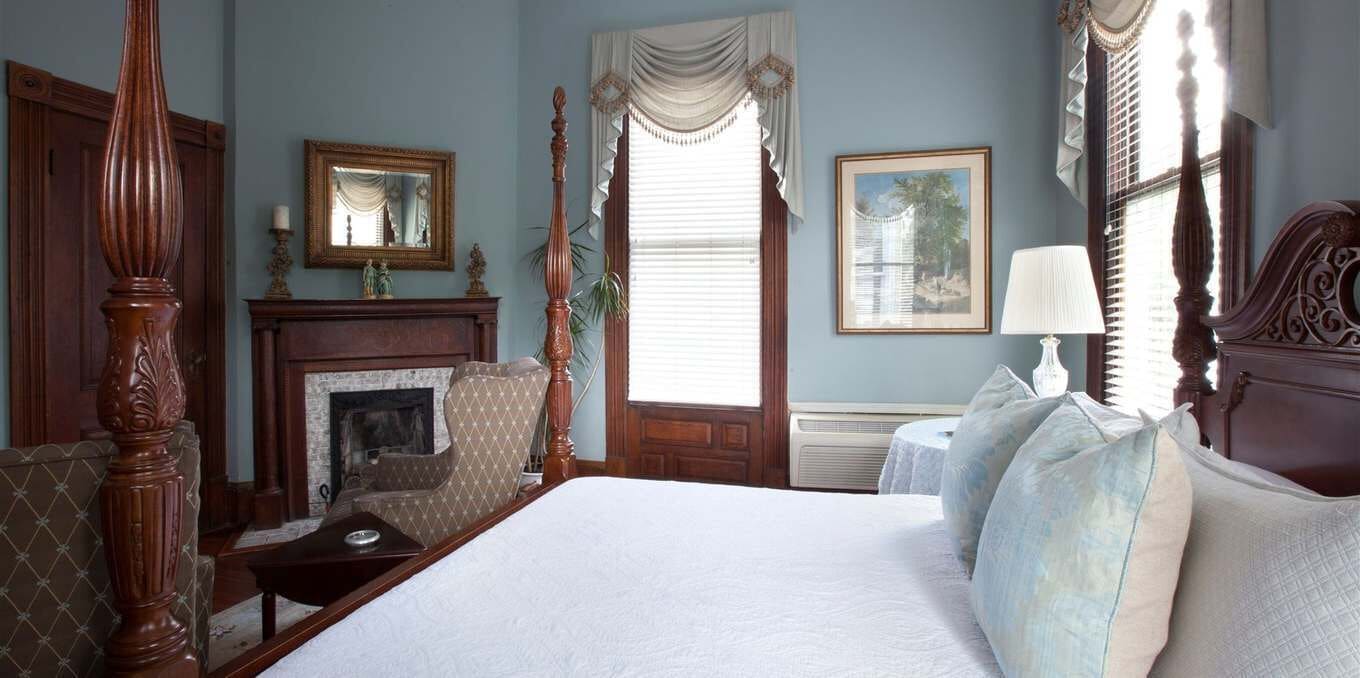 A bedroom with a four-poster bed, a fireplace, a large window with curtains, and a framed picture on the wall.