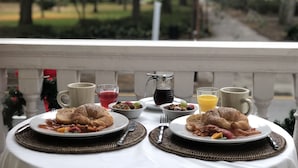 Una tavola imbandita per la colazione con croissant, frutta e bevande, su un balcone con vista su un parco.