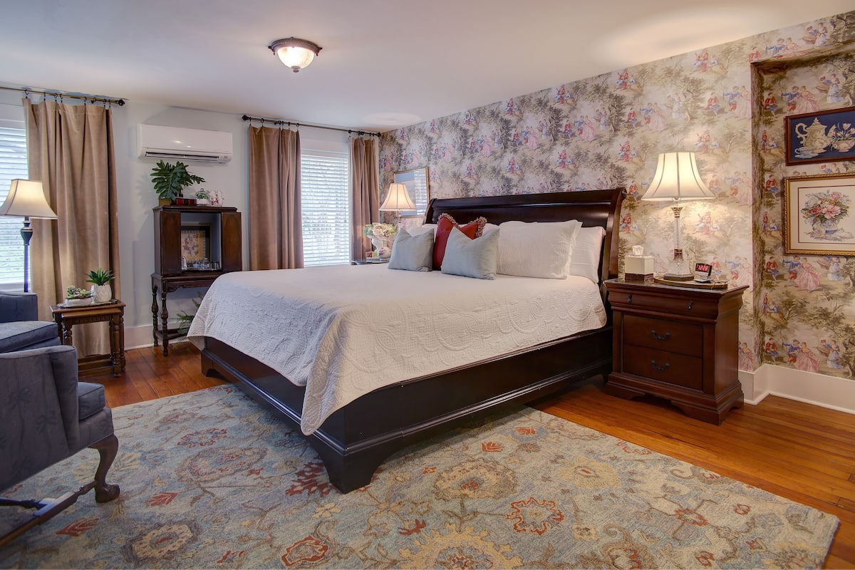 A neatly arranged bedroom with a large bed, a chair, a nightstand, and a wall with floral wallpaper.