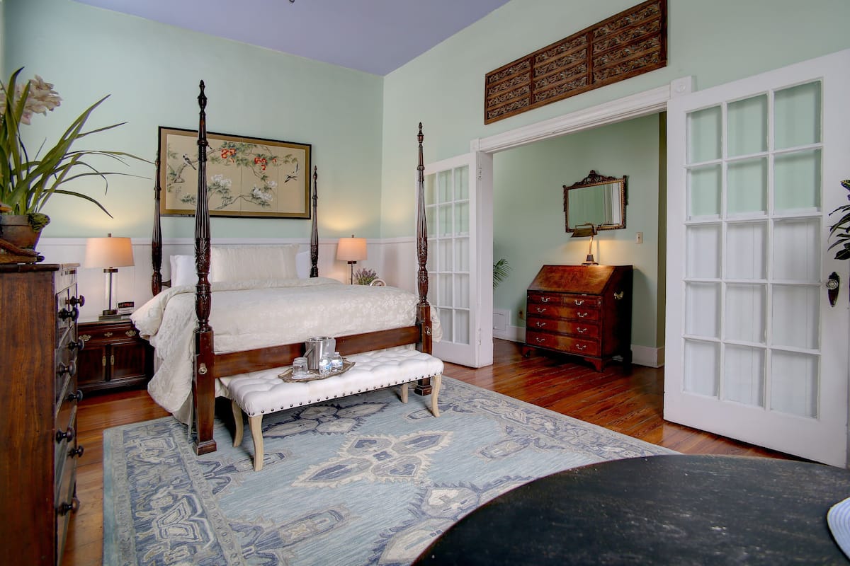 A four-poster bed with a bench, a dresser, and a mirror in a room with a large window and a door leading to another room.